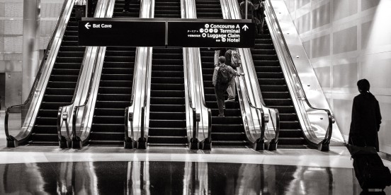 photo of an airport escalator system carrying passengers up while a sign to the side leads to the Bat Cave